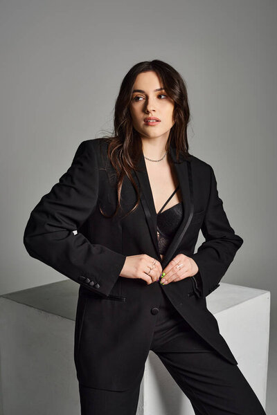 A beautiful plus size woman strikes a confident pose in a stylish black suit against a grey backdrop.