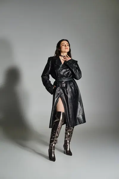 stock image A stylish plus size woman stands confidently in a black coat and boots against a gray backdrop, exuding elegance.