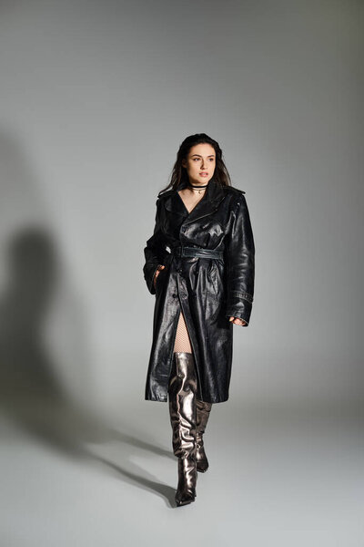 A stunning plus size woman strikes a pose in a sleek black coat and boots against a neutral backdrop.