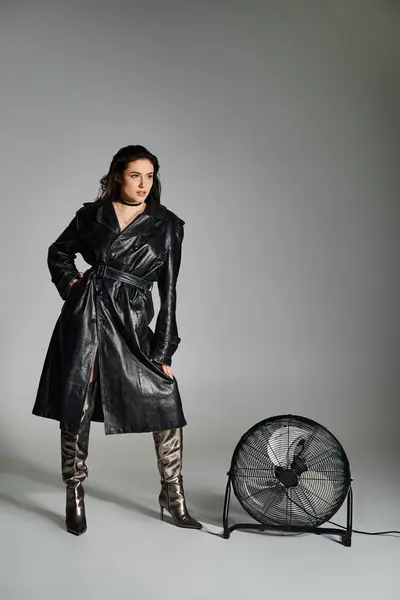 stock image Stunning plus size lady in chic black coat and boots standing gracefully next to a retro fan on gray background.