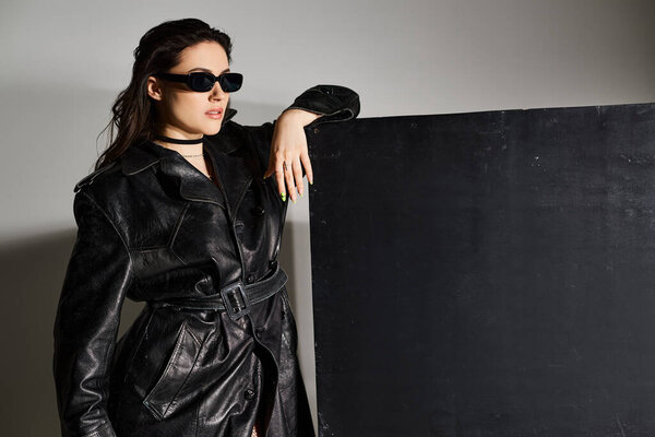 A stylish plus size woman strikes a pose in a black leather coat and sunglasses against a gray backdrop.