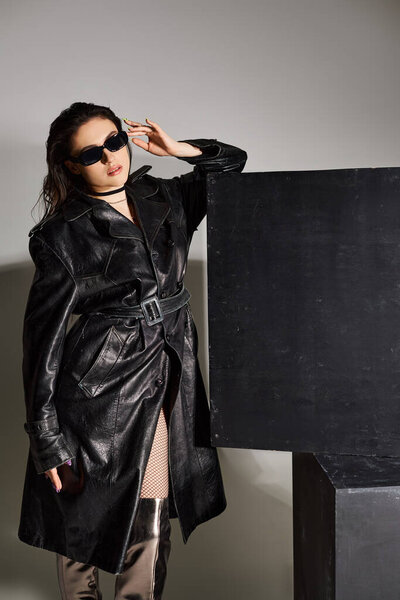 A stunning plus size woman strikes a pose in a black leather coat and thigh high boots on a gray backdrop.
