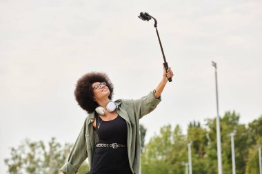 Kıvırcık saçlı genç bir Afrikalı Amerikalı kadın kaykay yaparken selfie çubuğu ve kulaklık takıyor açık hava kaykay parkında..