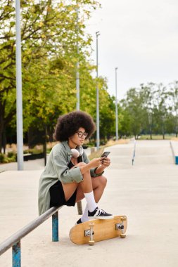 Kıvırcık saçlı genç Afrikalı Amerikalı bir kadın kaykay üzerinde oturuyor, açık hava kaykay parkında cep telefonuna sarılmış..