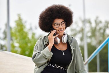 A stylish woman with an afro hairstyle chats on her smartphone while enjoying the outdoors. clipart