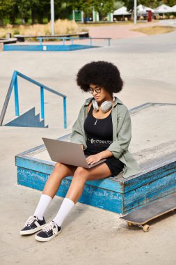 Kaykayın üzerinde oturan, kaykay parkında dizüstü bilgisayarda yazan kıvırcık saçlı genç bir Afrikalı Amerikalı kadın..