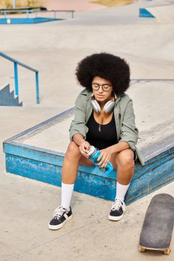 Kıvırcık saçlı genç bir Afrikalı Amerikalı kadın şehir kaykay parkında mavi bir kulübenin üstünde kaykayın yanında oturuyor..