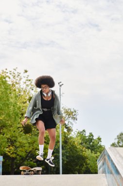 Kıvırcık saçlı genç Afrikalı Amerikalı bir kadın kaykayını açık hava kaykay parkında havaya fırlatıyor..
