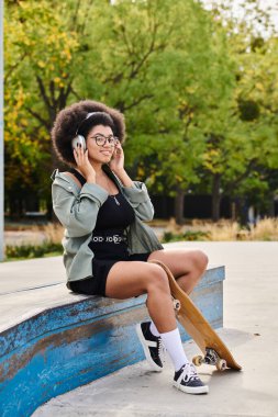 A young African American woman with curly hair sits on a bench, engrossed in a conversation on her cell phone at a skate park. clipart