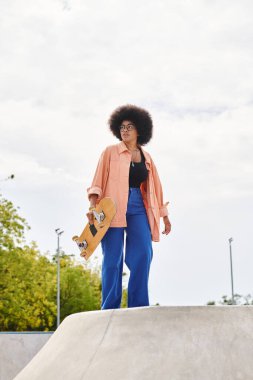 Kıvırcık saçlı, genç bir Afro-Amerikalı kadın açık hava kaykay parkında kaykay tutuyor..