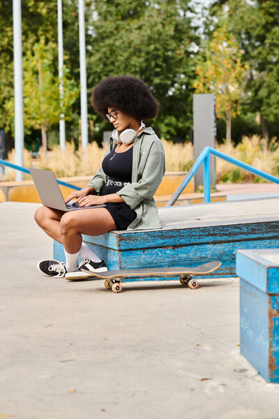 A young African American woman with curly hair sits on a bench outdoors, intensely working on her laptop.