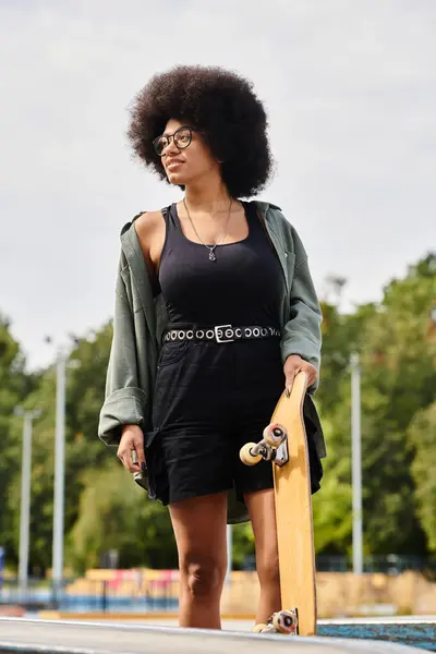 stock image A young African American woman confidently holds a skateboard on top of a ramp in a skate park.