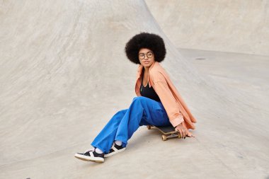 Kıvırcık saçlı Afro-Amerikan bir kadın canlı bir kaykay parkında kaykay üzerinde oturuyor..