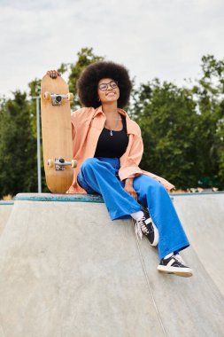 Kıvırcık saçlı genç bir Afrikalı Amerikalı kadın kaykay rampasının tepesinde oturuyor. Kendine güveni ve azmi var..