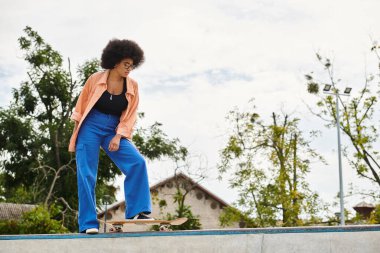 Young African American woman with curly hair riding skateboard on top of cement wall in outdoor skate park. clipart