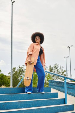 A young African American woman with curly hair confidently stands on steps, holding a skateboard in an urban skate park. clipart