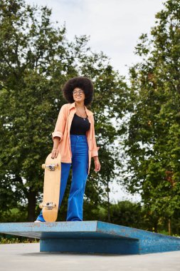 Kıvırcık saçlı genç Afrikalı Amerikalı kadın kaykay parkında mavi bir platformun üstünde kaykay tutuyor..