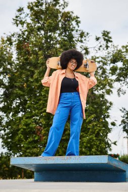 Kıvırcık saçlı Afrikalı Amerikalı bir kadın kaykay parkında mavi bir platformda kaykay tutarken poz veriyor..