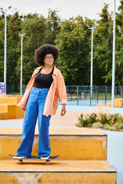 A young African American woman with curly hair confidently stands atop a skateboard ramp in an outdoor skate park.