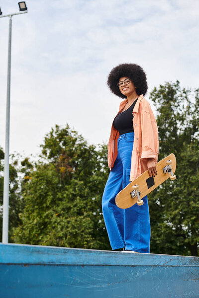Young African American woman with curly hair confidently holds skateboard, poised on ramp in skate park.