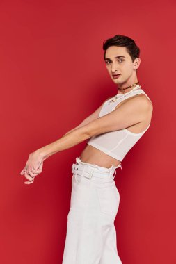 attractive gay man in casual attire with accessories posing on red background and looking at camera clipart