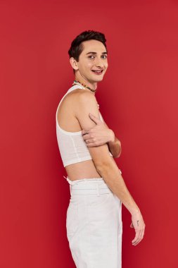 Beyaz giysili şık aksesuarları olan neşeli yakışıklı eşcinsel adam kırmızı arka planda kameraya bakıyor.
