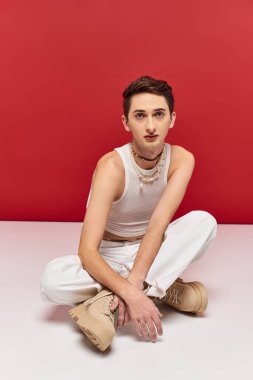 good looking androgynous man in casual attire sitting on floor and looking at camera on red backdrop clipart