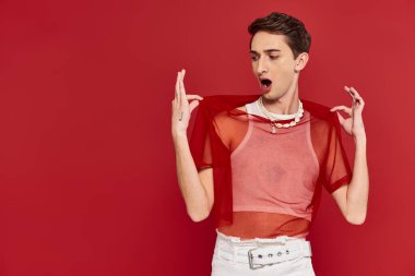 handsome shocked androgynous man with red stylish fishnet posing emotionally on red backdrop clipart