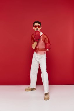 Şok olmuş güneş gözlüklü, kırmızı balık ağlı boks eldivenli ve kameraya bakan gay adam.