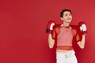 Kırmızı arka planda boks eldiveni takmış sıradan giyinmiş neşeli, iyi giyimli gay adam kameraya bakıyor.