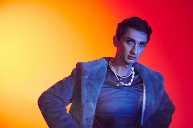 sofistike, çift cinsiyetli, mor kürklü bir adam canlı arkaplanda poz veriyor ve kameraya bakıyor.