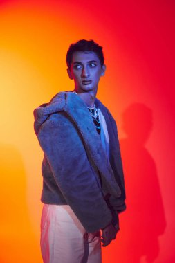 Güzel görünümlü, çift cinsiyetli, mor kürklü, canlı arka planda poz veren ve başka yöne bakan bir adam.