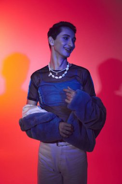 Neşeli, çift cinsiyetli, mor sahte kürklü bir adam canlı arka planda poz veriyor ve başka tarafa bakıyor.