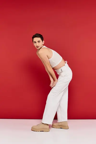 stock image fashionable gay man in casual attire with accessories posing on red background and looking at camera