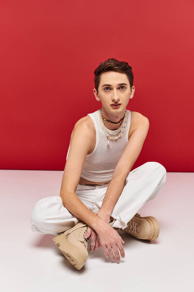 good looking androgynous man in casual attire sitting on floor and looking at camera on red backdrop