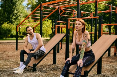 Bir erkek ve bir kadın, spor kıyafetleriyle, parkta banklarda otururlar, fitness seansına katılırlar.