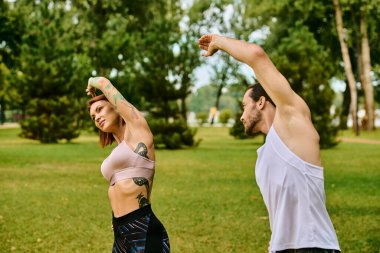Spor kıyafetleriyle yoga yapan bir erkek ve bir kadın huzurlu bir park ortamında birlikte poz veriyorlar.