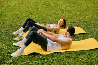Spor kıyafetli bir adam ve bir kadın çimlerin üzerinde oturmuş yoga pozları veriyorlar.