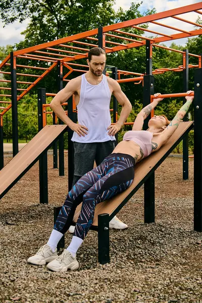 stock image A determined man and woman in sportswear stand confidently on a wooden bench during a challenging outdoor workout session.