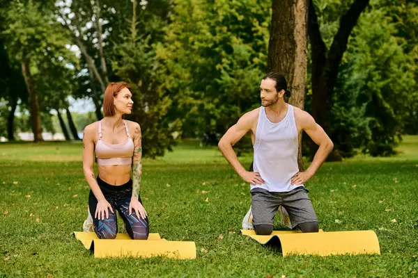 stock image A determined man and woman in sportswear sit in the grass, guided by a personal trainer during a workout session.
