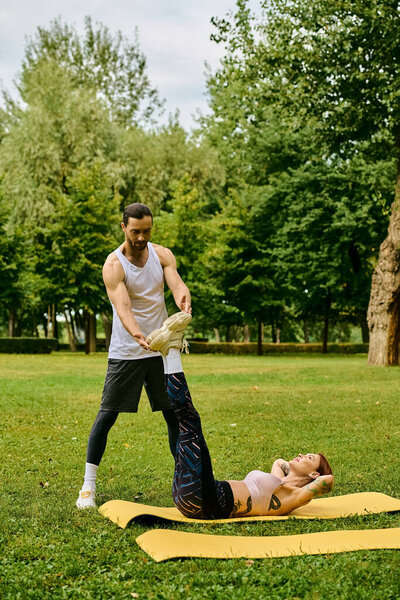 A determined man stands over a woman as they exercise on a yellow mat outdoors. The personal trainer motivates and guides the woman.