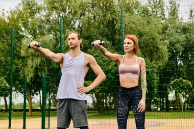 Bir erkek ve bir kadın, spor kıyafetleri içinde, dambıllarla egzersiz yapıyor, birlikte egzersiz yaparken azim ve odaklanma gösteriyor.