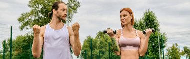 Spor giyim sektöründe erkek ve kadın birlikte ayakta, halterlerle egzersiz yapıyorlar.