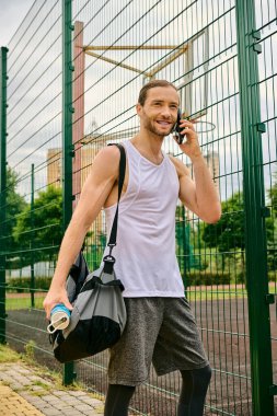 A man in casual attire stands beside a fence having a conversation on his cell phone. clipart
