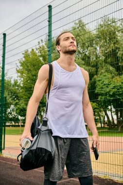 Spor giyimli kararlı bir adam tenis kortunda elinde bir çantayla dolaşıyor.