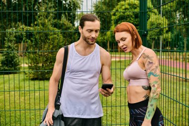 Spor kıyafetleri içinde kararlı bir erkek ve kadın akıllı telefon kullanarak çitin önünde dikiliyorlar.
