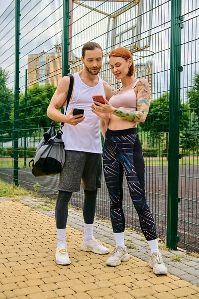 stock image A man and woman in sportswear stand side by side outdoors, holding smartphones