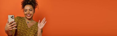 A beautiful African American woman in stylish attire holding a cell phone in front of an orange wall. clipart