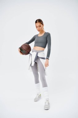 Genç ve sportif bir kadın, gri bir arka plana karşı etkin bir şekilde giyinmiş bir basketbol topunu nazikçe sağ elinde tutuyor..