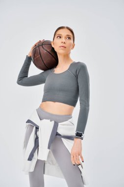 Sportif genç bir kadın, gri bir arka planda atletik hünerini sergileyerek, sağ elinde bir basketbol topu tutuyor..